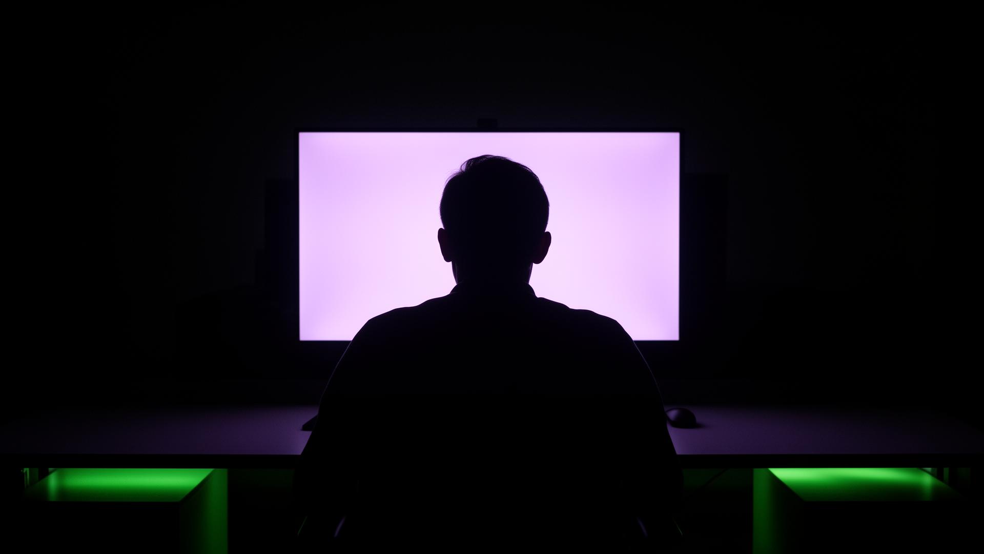 Silhueta de uma pessoa de costas diante de um monitor brilhando em violeta, escritório escuro